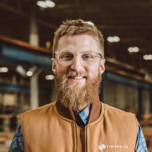 A mean wears a plaid shirt under a zip-up vest. He has short hair, a beard, and glasses. He smiles at the camera.