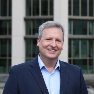 A man wears a collared shirt and a suit jacket. He stands in front of a building and smiles at the camera.