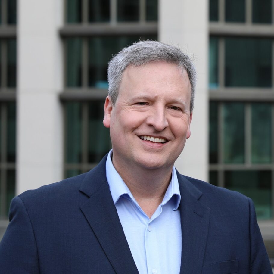 A man wears a collared shirt and a suit jacket. He stands in front of a building and smiles at the camera.