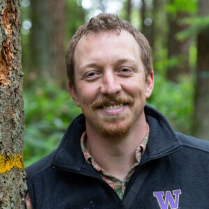 A man with curly, close cropped hair wears a collared shirt under a quarter-zip jacket. He leans against a tree and smiles at the camera.