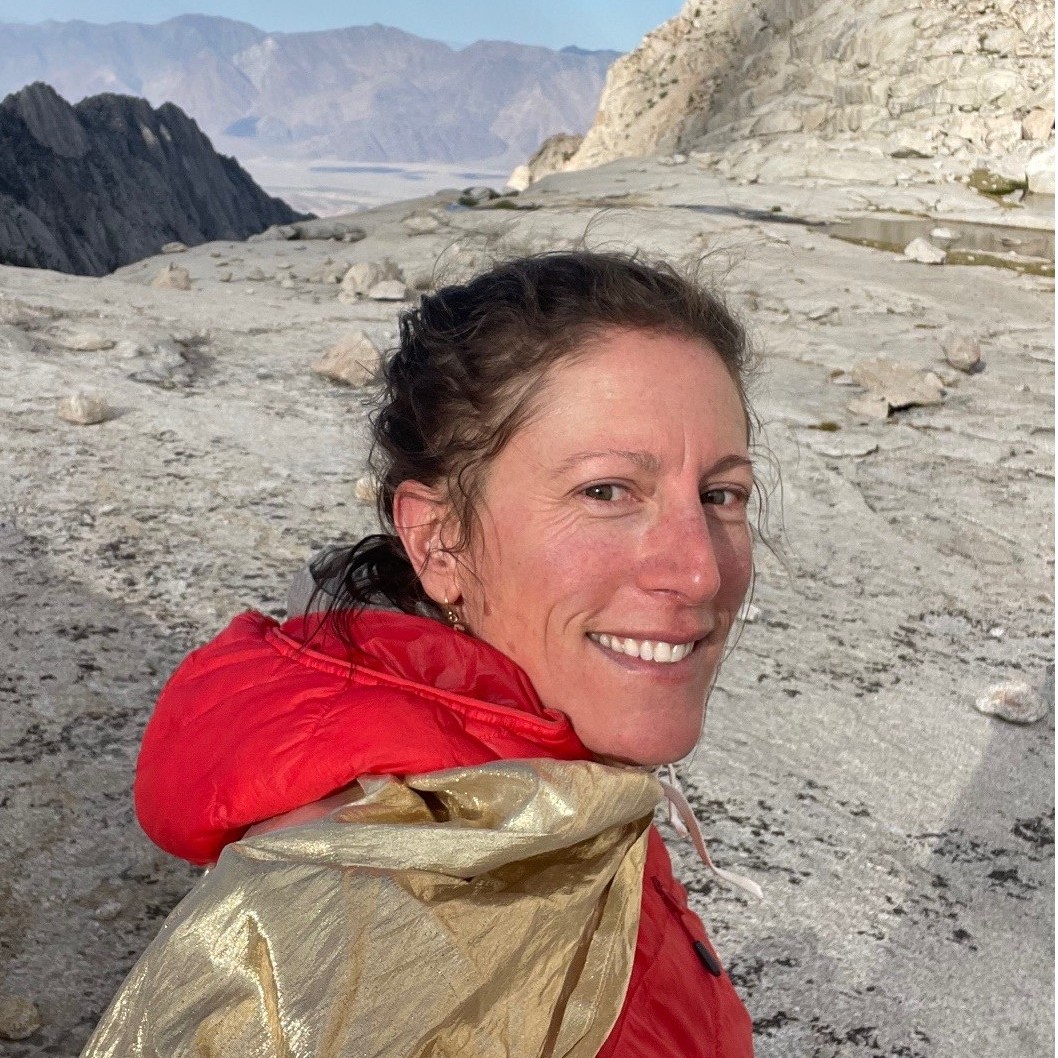 A woman wears a hooded jacket and stands with her side towards the camera. She turns her face and smiles at the camera. In the background is a rocky, mountainous landscape.