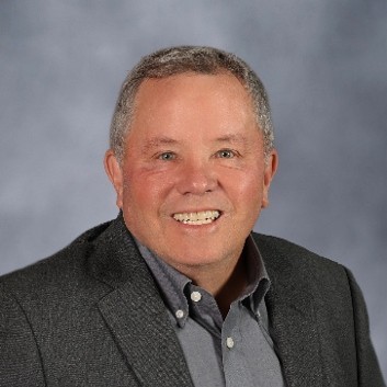 A man wears a collared shirt and suit jacket. He smiles at the camera.